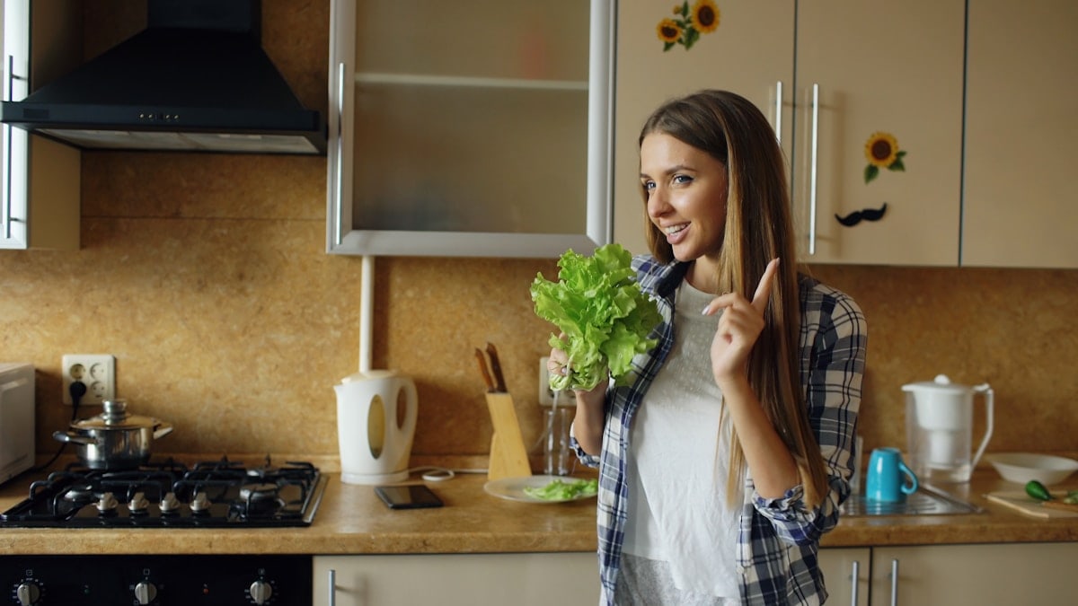 Cheerful young funny woman dancing and singing with lettuce microphone while cooking breakfast in the kitchen early morning at home